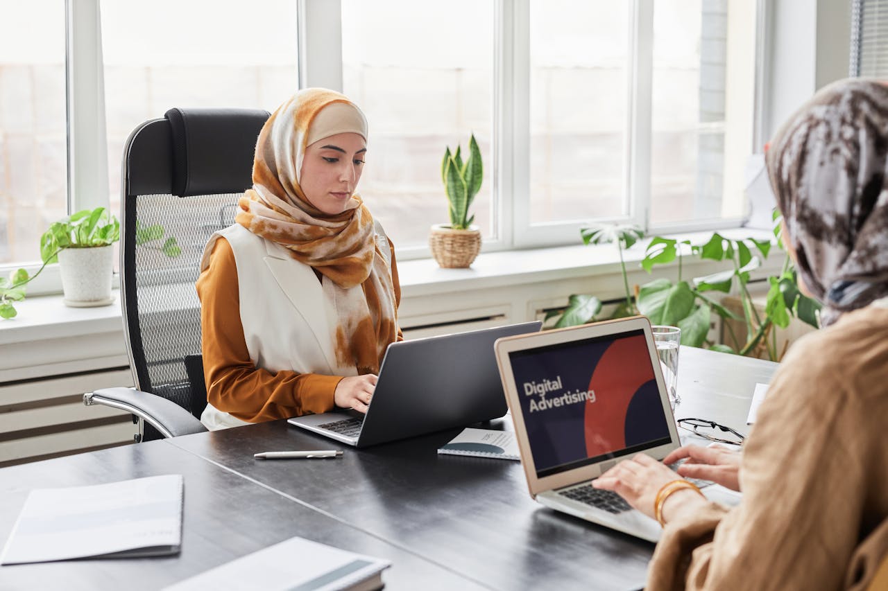Two women in hijabs working on laptops in a bright, modern office.